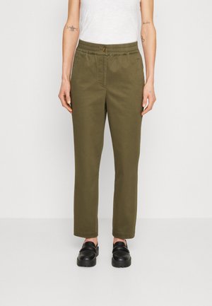 Kobieta nosząca oliwkowo zielone spodnie o prostych nogawkach z guzikami i elastycznym paskiem, w połączeniu z czarnymi mokasynami i białym bezrękawnikiem.