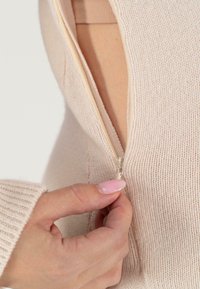 Pull en maille beige clair avec une fermeture éclair à moitié ouverte. La texture du tissu est douce et côtelée, avec des coutures visibles et une coupe ajustée.
