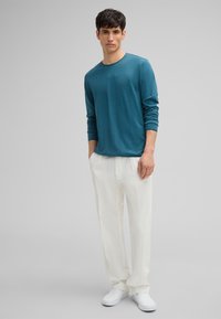 Teal Langarmshirt mit rundem Halsausschnitt, kombiniert mit weißen, lockeren Hosen und weißen Slippern. Einfaches, lässiges Design.