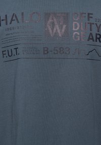 Niebieska tkanina z wyblakłym czarnym tekstem i grafiką, w tym napisami "HALO", "OFF DUTY GEAR" oraz "PHYSICAL EDUCATION & TRAINING" z abstrakcyjnym zarysem gór.