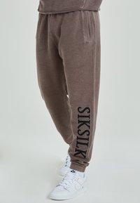 Bruna mjukisbyxor i texturerad tyg, elastisk midja, sidofickor och en svart "SIKSILK"-logotyp längs benet. Matchas med vita sneakers.