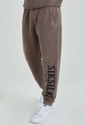 Pantalons de jogging marron en tissu texturé, taille élastique, poches latérales et logo noir "SIKSILK" le long de la jambe. Assortis à des baskets blanches.