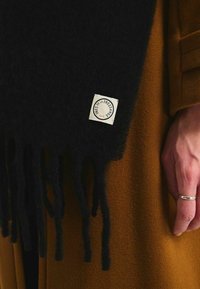 Écharpe en laine noire avec des bords frangés et une étiquette blanche comprenant un logo circulaire. Portée par-dessus un vêtement texturé marron.