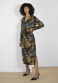 Robe à fleurs en tissu foncé avec des motifs jaunes et bleus, dotée d'une ceinture à nouer, de manches longues et de fentes sur les côtés. Associée à des talons noirs.