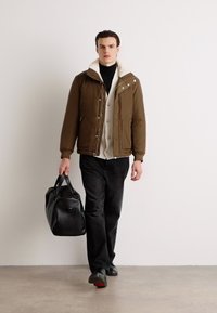 Brun isoleret jakke med shearlingkrave, beige skjorte med knapper, sorte bukser og bærer en sort læder duffeltaske.