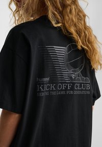 Melna kokvilnas T-kreklu ar pelēku grafiku volejbola un tekstu: "hummel KICK OFF CLUB" ar svītrotiem akcentiem aizmugurē.