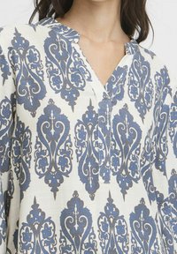 Blouse en tissu léger à motifs avec un fond blanc et des dessins bleus complexes. Dotée d'un col en V et d'un détail à boutons sur le col.