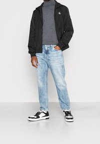 Veste zippée noire par-dessus un col roulé gris, jeans bleu clair délavés, et baskets noires et blanches, avec une coupe décontractée et un style casual.