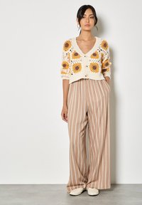 Cárdigan de crochet con girasoles en crema, combinado con pantalones de pierna ancha a rayas en marrón claro. Tejido texturizado con cierre de botones, zapatos planos.