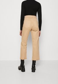 Pantalones de pierna ancha beige de cintura alta con dobladillo deshilachado, con dos bolsillos traseros, combinados con botas negras de tacón y una blusa negra ajustada.