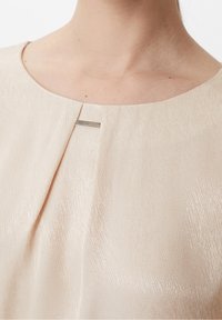 Nærbillede af en beige tekstureret top med rund halsudskæring og en vandret sølvstænger detalje nær kravebenet.