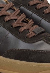 Brązowe zamszowe i gładkie czarne skórzane sneakersy z brązowymi sznurowadłami oraz gumową podeszwą. Zauważalny detal logo na uchwycie sznurowadeł.