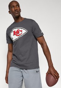 Grå kortærmet t-shirt med et stort rødt og hvidt Kansas City Chiefs-logo; iført lysegrå atletikshorts; holder en fodbold.