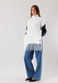 Poncho bianco oversize con collo alto, rifinito con frange bianche. Abbinato a jeans a gamba larga blu e scarpe nere con punta appuntita.