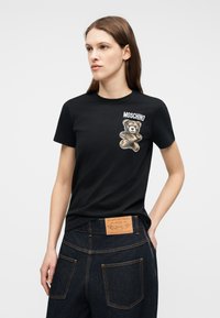 Mujer con camiseta negra de Moschino con gráfico de oso de peluche en el pecho y vaqueros de denim oscuro con etiqueta de parches de cuero marrón.