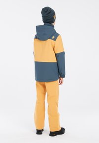 Wasserfeste Jacke in Blau und Gold mit Kapuze, kombiniert mit gelben Hosen und schwarzen Stiefeln. Mit elastischen Bündchen und minimalem Branding.