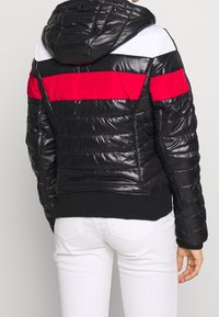 Vadderad svart jacka med röda och vita horisontella ränder, med huva och ribbad mudd, som visar en glänsande, texturerad yta.