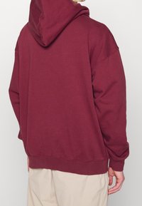 Osoba oblečená v burgundské mikině s kapucí a béžových kalhotách, zobrazená zezadu na jednoduchém světle šedém pozadí.