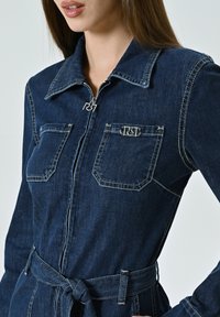 Dunkelblauer Jeans-Overall mit einem Reißverschluss vorne, Punktkragen, zwei Brusttaschen und einem Gürtel an der Taille. Mit silberfarbenen Hardware-Details.