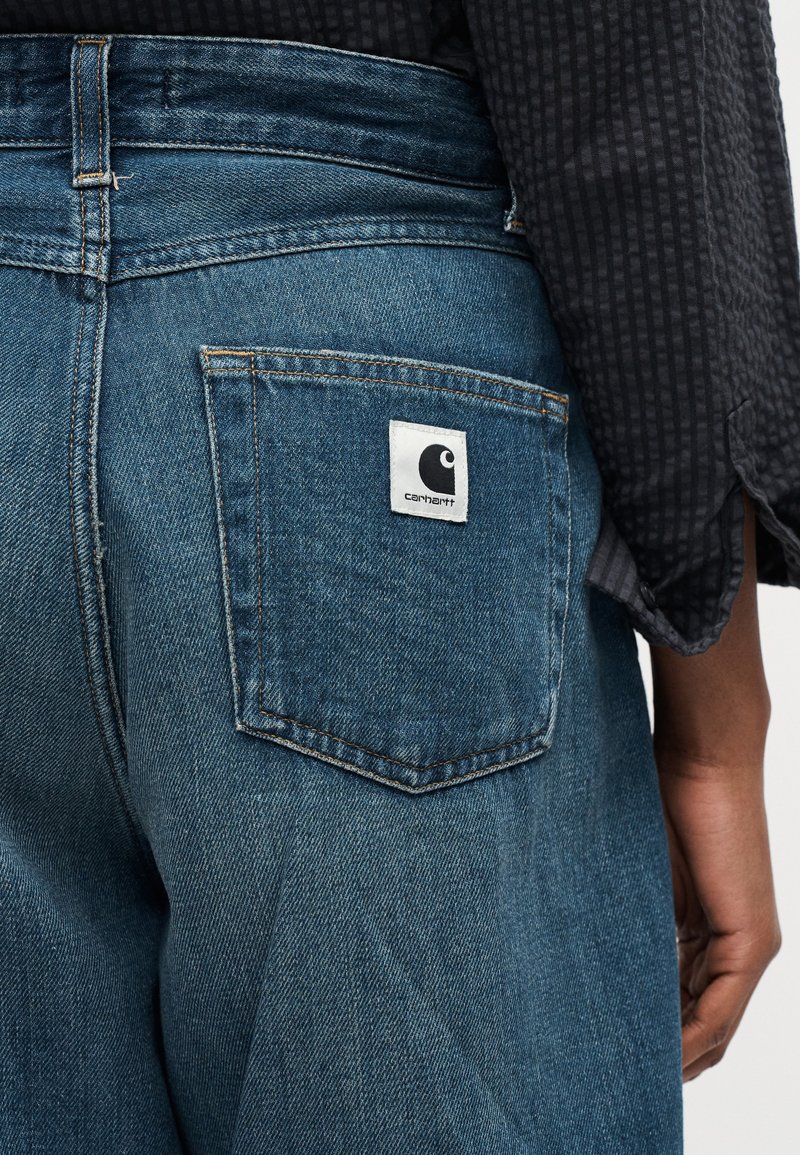 Primo piano della tasca posteriore di jeans di denim blu con toppa del logo Carhartt, indossati da persona con camicia a maniche lunghe scura e strutturata.