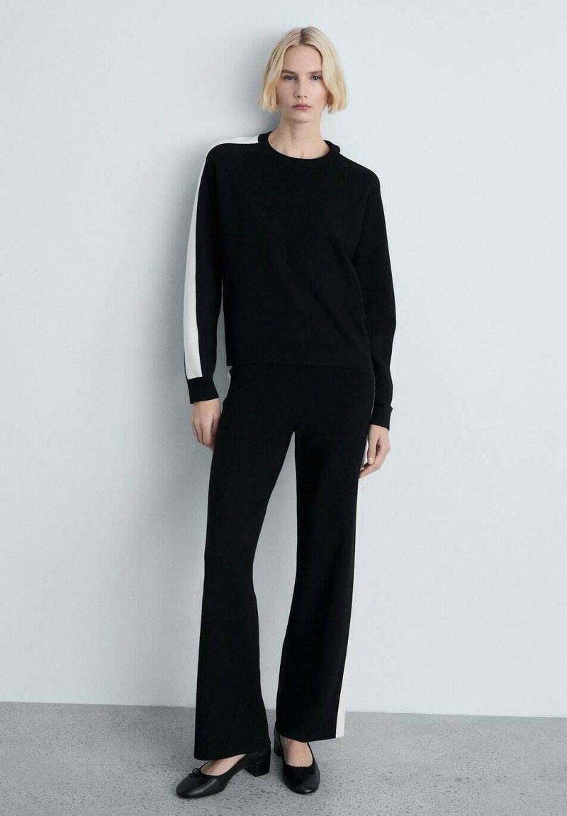 Zwarte sweatshirt met een witte accent op één mouw, gecombineerd met zwarte wijde broek. Gladde textuur; minimalistisch ontwerp. Zwarte schoenen.