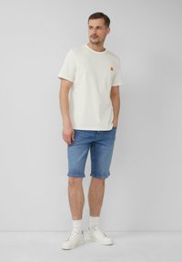 Weißes Baumwoll-T-Shirt mit einem kleinen orangen Flammenlogo, kombiniert mit knielangen blauen Jeansshorts und weißen Sneakern mit grauen Akzenten.