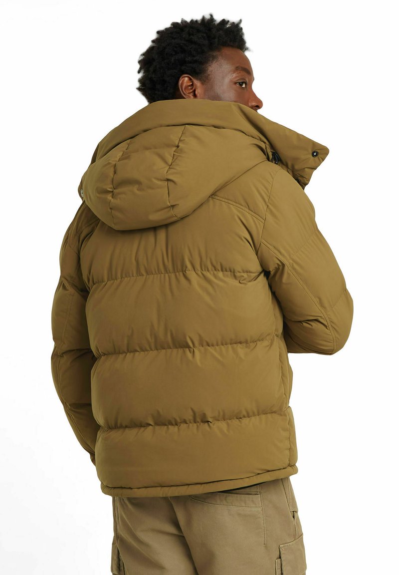 Brązowa ocieplana kurtka z kapturem, z pikowanym wzorem i zapięciem na zamek błyskawiczny, noszona na jasnych spodniach cargo.