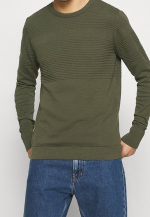 Muž v olivově zeleném pleteném svetru s dlouhými rukávy a modrých džínách, stojící s rukama blízko kapes.