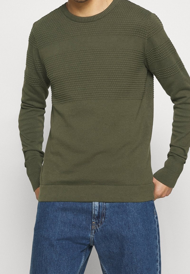 Homme portant un pull en maille texturée vert olive à manches longues et un jean bleu, debout les mains près des poches.