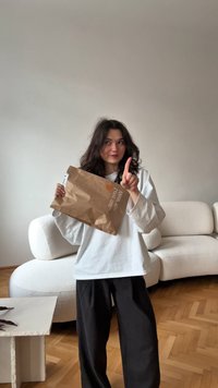 Brun papirpose med et hjerte-logo, holdt af en person iført en løs hvid skjorte og sorte bukser, stående i et minimalistisk rum.