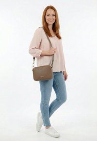 Glimlachende vrouw met rood haar, lichtroze blouse, blauwe jeans en witte sneakers, met een bruine schoudertas.