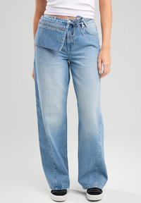 Jeans de mezclilla de pernera ancha en azul claro con un bolsillo frontal con cremallera y cintura con cordón. Textura suave con un acabado desgastado y estilo informal.