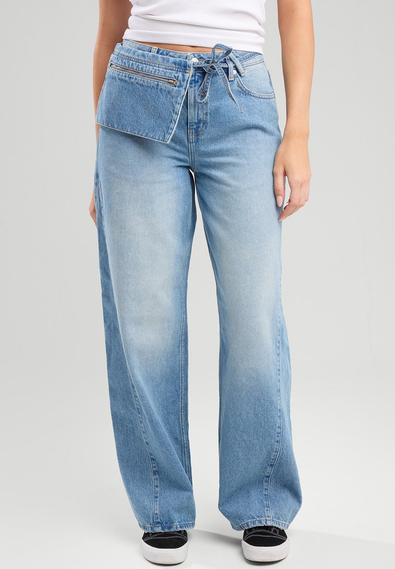 Jeans de mezclilla de pernera ancha en azul claro con un bolsillo frontal con cremallera y cintura con cordón. Textura suave con un acabado desgastado y estilo informal.