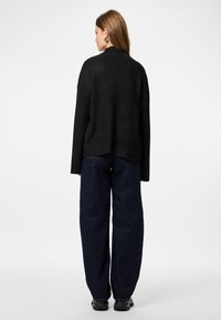 Sieviete ar gariem brūniem matiem, tērpusies melnā ribotā džemperī, tumšos platākajos džinsos un melnās sporta kurpēs, stāv pretējo virzienā uz vienkārša fona.