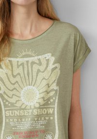 T-shirt vert olive avec un col rond et des manches courtes retroussées, mettant en avant un design graphique jaune clair représentant des rayons de soleil et du texte.