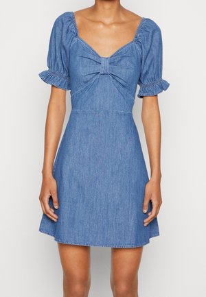 Kvinde iført en kort blå denimkjole med puffede korte ærmer, en firkantet udskæring og en knudedetalje ved brystet.
