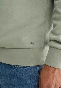 Sudadera verde claro hecha de tela texturizada, con puños y dobladillo acanalados, y un pequeño detalle de logo cerca de la parte inferior. Llevada con denim.