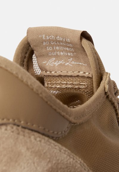 Καφέ αθλητικό παπούτσι με υλικά σουέτ και δίχτυ. Η γλώσσα του παπουτσιού φέρει ένα απόσπασμα και το όνομα της μάρκας σε λευκό, με ορατές ραφές στα πλαϊνά.