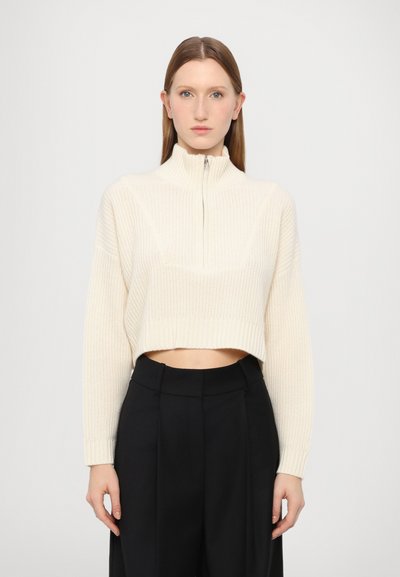 Cremefarvet cropped sweater med høj krave, ribbet tekstur og lynlåslukning, parret med sorte højtaljede bukser.