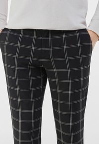 Černé kalhoty s mřížkovým vzorem, s bílými liniemi. Vyrobené z hladké látky, mají střih na míru a boční kapsy.