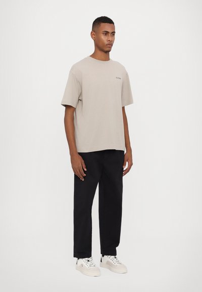 Beige bomulls T-skjorte med avslappet passform, korte ermer og en liten logo aksent. Kombinert med svarte bukser og hvite joggesko.