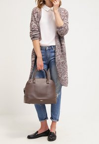 Donna che indossa un polo bianco, un lungo cardigan bordeaux, jeans blu arrotolati e mocassini neri, mentre tiene una grande borsa in pelle marrone per le maniglie.