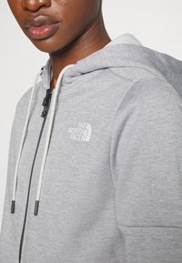 Gros plan d'une personne portant un sweat à capuche zippé gris clair avec des cordons blancs et le logo The North Face sur la poitrine.