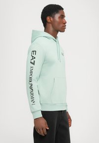 Light mint green hoodie with a front pocket, ribbed cuffs, and black branding printed down the left sleeve. Soft fabric texture.