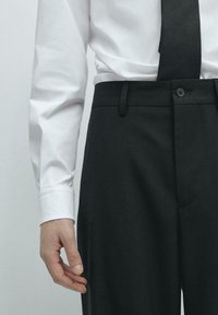 Schwarze, maßgeschneiderte Hose mit glatter Textur, ausgestattet mit einer Fronttasche, Knopfverschluss und schmaler Passform, kombiniert mit einem weißen Hemd.