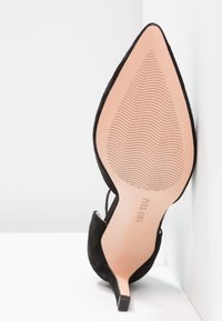 Chaussure à talon haut noire à bout pointu inclinée vers le haut, montrant une semelle lisse avec un motif ondulé antidérapant et le nom de la marque "Pura One".