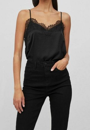 Vrouw draagt een zwarte satijnen camisole met kanten randje en dunne bandjes en hoge zwarte jeans, hand in zak tegen een effen achtergrond.