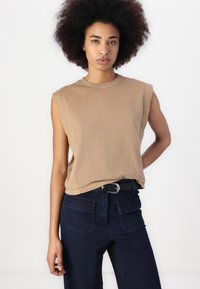 Top sin mangas de color bronceado, hecho de una tela suave, combinado con pantalones vaqueros oscuros de tiro alto y un cinturón negro con una hebilla plateada.