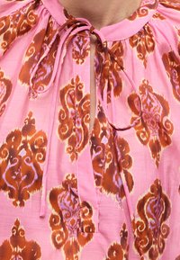 Gros plan sur un chemisier rose avec un motif floral marron et lavande, avec un col noué en nœud papillon et des détails de tissu froncé.