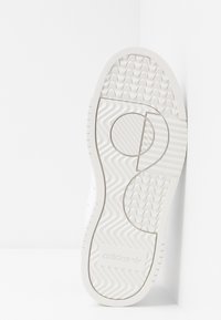 Bílá gumová podrážka tenisky s texturovanými gripy, vybavená kruhovým designovým prvkem a reliéfním logem. Hladký povrch s jasnými vzory.